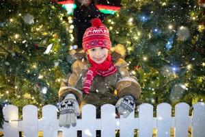 lindo criança em Natal rua. uma Garoto dentro inverno roupas em a fundo do Natal árvores e neve. foto