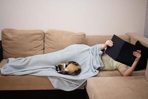 jovem mulher deitado dentro cama às casa e ler livro com dela gato. foto