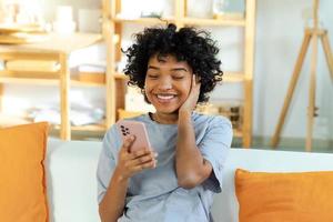 animado feliz jovem Preto africano americano mulher segurando célula telefone rindo sentindo-me alegria obtendo Móvel mensagem. muito feliz menina rindo em voz alta sentado em sofá assistindo engraçado vídeo lendo notícias. foto