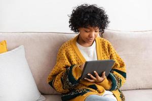 garota afro-americana segurando a tela de toque do tablet digital digitando a página de rolagem em casa. mulher com dispositivo de tecnologia móvel navegando na internet usando aplicativos de mídia social jogando jogo. estudar, trabalhar, fazer compras online. foto