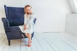 doce pequeno menina dentro branco vestir sentado em moderno acolhedor azul cadeira relaxante dentro branco brilhante vivo quarto às casa dentro de casa. infância escolares juventude relaxar conceito. foto