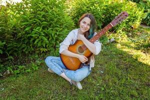 jovem mulher sentado dentro Relva e jogando guitarra foto