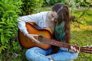 jovem mulher sentado dentro Relva e jogando guitarra foto