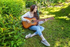 jovem mulher sentado dentro Relva e jogando guitarra foto