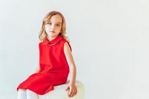 doce pequeno menina dentro vermelho vestir sentado em cadeira contra branco parede às lar, relaxante dentro branco brilhante vivo quarto dentro de casa. infância escolares juventude relaxar conceito. foto