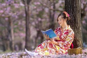 japonês mulher dentro tradicional quimono vestir sentado debaixo cereja Flor árvore enquanto lendo uma livro durante Primavera sakura festival conceito foto