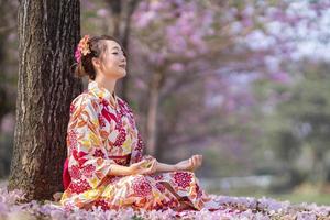 japonês mulher dentro quimono vestir é fazendo meditação debaixo sakura árvore durante cereja florescendo estação para interior paz, atenção e zen prática conceito foto