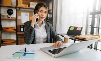 mulher asiática de negócios falando ao telefone e usando um laptop com um sorriso enquanto está sentado no escritório foto
