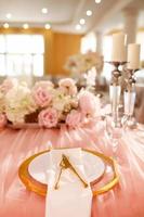 decoração de mesa com toalha de mesa rosa, castiçais de cristal com velas e flores rosa brancas no restaurante. dia de casamento elegante. mesa de ajuste com placa de ouro, garfo e faca foto