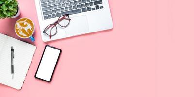 mesa de escritório com computador portátil, telefone inteligente de tela em branco, xícara de café, caneta, caderno em fundo rosa, vista superior com espaço de cópia, mock up. foto