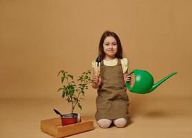 adorável criança menina detém rega pode e jardim ferramentas, sentado de de madeira engradado com florescendo Pimenta plantar dentro flor Panela foto