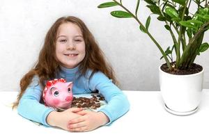 pequeno menina às a mesa alegremente abraços uma porquinho banco e dinheiro, poupança, financeiro alfabetização foto