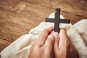 conceito do cristão. fechar acima do mão Rezar segurando uma Jesus Cristo Preto Cruz em a velho de madeira mesa fundo com cópia de espaço foto