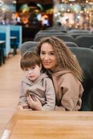 retrato do uma lindo jovem mãe e uma criança de quatro anos filho dentro dela braços. a família é sentado dentro uma cafeteria em uma cadeira foto