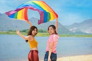 feliz lésbica, lindo ásia jovem dois mulheres, menina gay, casal amor momento gastos Boa Tempo junto, segurando ou acenando lgbt arco-íris, orgulho bandeira juntos em a de praia. foto