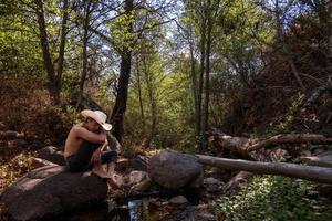 homem senta em uma Rocha dentro uma floresta, vestindo uma vaqueiro chapéu. foto