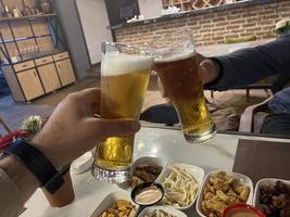 mãos do homens tilintar Cerveja óculos com lanches em a mesa foto