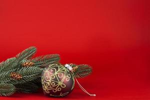 Natal cartão. verde galhos do uma Natal árvore com cones e uma Natal bola com ouro enfeites em uma vermelho fundo foto