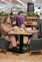jovem encaracolado cabelos mãe e dela pequeno filho ter café da manhã ou almoço dentro uma cafeteria, sentado às uma mesa foto
