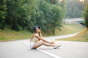 uma feliz menina dentro branco calção e uma amarelo blusa detém uma vidro do suco com uma Palha dentro dela mãos e senta em a estrada perto a floresta foto