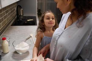 lindo feliz criança, adorável pequeno menina sorrisos fofinho olhando às dela amoroso mãe, em pé às cozinha bancada e cozinhando junto, preparando massa para a comemorar encolher terça foto