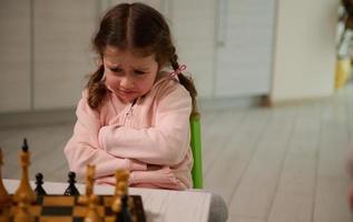 uma pequeno frustrado menina senta dentro a cadeira com cruzado braços e a ofendido olhar, parece às a tabuleiro de xadrez enquanto jogando com dela irmão, ensino dela a jogos foto