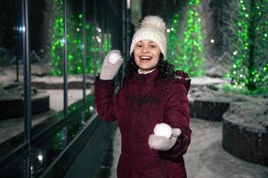 bonita mulher tocam com bolas de neve em inverno Nevado noite, em rua iluminado de guirlandas. inverno Diversão - bola de neve luta foto