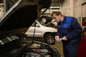 jovem profissional mecânico escreve em prancheta enquanto inspecionando a motor do a automóvel sagacidade aberto de capuz dentro auto serviço reparar fazer compras foto