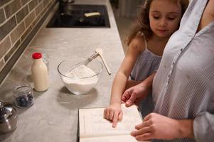 lindo feliz criança, adorável pequeno menina pontos com dela dedo em uma receita livro, em pé às cozinha bancada fechar para dela mãe enquanto desfrutando cozinhando junto, preparando massa para encolher terça foto