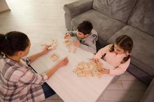 a sobrecarga Visão do uma mãe e crianças jogando borda jogos, construção de madeira estruturas, construções em a mesa dentro casa vivo sala. família passatempo e educacional lazer conceito foto