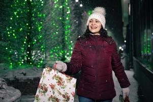 bonita mulher caminhando com compras bolsas em a rua iluminado de guirlandas às Nevado inverno noite foto