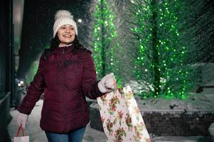 delicioso hispânico mulher anda em com compras bolsas em uma cidade rua iluminado de guirlandas às Nevado inverno noite foto