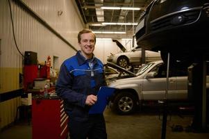 profissional retrato do uma jovem caucasiano auto engenheiro técnico mecânico dentro uniforme sorridente com uma cheio de dentes sorrir olhando às Câmera enquanto em pé dentro reparar fazer compras com prancheta dentro dele mãos foto