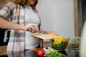 grávida mulher segurando uma corte borda e derramando picado amarelo Sino Pimenta a partir de isto para dentro uma tigela com uma saudável vegetal salada, preparando uma vegetariano almoço enquanto em pé às a cozinha ilha foto