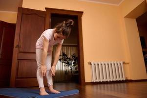 adorável ativo pequeno criança menina exercício , trabalhando fora, praticando ioga, em pé em ginástica esteira dentro acolhedor casa interior foto