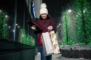 mulher com compras bolsas e Natal presentes, caminhando baixa a rua, iluminado de iluminação às inverno Nevado noite. foto