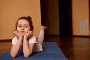 encantador criança menina deitado em barriga em uma ginástica esteira, olhando às Câmera. ioga prática. fitness. infância. esporte conceito foto