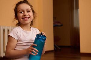 retrato do 5-6 anos velho positivo desportivo criança menina segurando garrafa do água, sorridente uma cheio de dentes sorrir olhando às Câmera foto