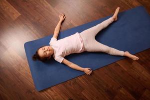 topo Visão pequeno menina relaxante, deitado em uma azul ginástica esteira em de madeira chão depois de praticando ioga às casa ou ioga estúdio foto