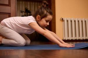 fechar-se pequeno menina alongamento dela corpo dentro balasana pose, sentado dentro criança pose enquanto praticando ioga em uma ginástica esteira foto