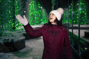 delicioso mulher pega flocos de neve, goza caminhando baixa a cidade rua, iluminado de guirlandas em uma Nevado inverno noite foto