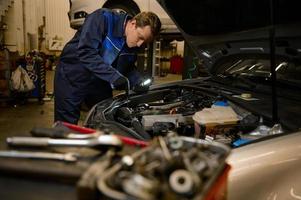 jovem concentrado garagem mecânico, técnico, carro engenheiro segurando uma lanterna luminária e reparação carro debaixo de capuz dentro a reparar fazer compras. auto serviço, trabalho, pessoas conceito foto