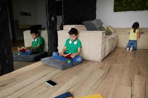 adorável diverso caucasiano crianças dentro a crianças sala. adorável Adolescência escola Garoto lendo livro, enquanto sentado às a sofá dentro dele quarto em a fundo do dele mais jovem irmã jogando jogos foto