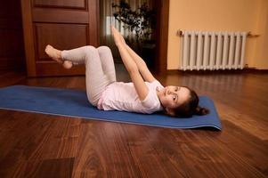 feliz relaxado caucasiano pequeno criança menina levantando braços, fazendo abdômen exercícios, alongamento corpo, deitado em ginástica esteira foto