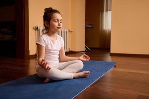 adorável bebê menina dentro branco camiseta e cinzento leggins, sentado dentro lótus pose em ginástica , meditando às casa interior foto