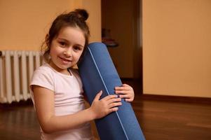 fechar-se retrato do uma sorridente pequeno menina segurando ginástica esteira, olhando às Câmera, desfrutando exercite-se e ginástica dentro de casa foto