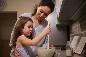 adorável pequeno menina tendo diversão, concentrado em mistura ingredientes e amassar massa dentro uma vidro tigela enquanto dela amoroso feliz mãe ensino dela para cozinhar panqueca massa. encolher terça conceito foto