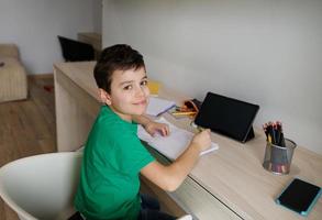 bonito caucasiano 9 anos velho escola Garoto senta às de madeira escrivaninha dentro frente do uma digital gadget e fazendo dele trabalho de casa, fofinho sorrisos olhando às Câmera. Educação e escola conceito. conectados Aprendendo foto