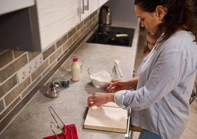 lindo mulher, dona de casa procurando uma receita em uma livro, em pé com dela filha às cozinha bancada com cozinhando utensílios e ingredientes para cozimento massa foto