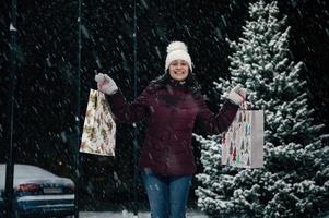 lindo africano americano mulher dentro caloroso roupas, com compras bolsas sorridente para a Câmera em uma Nevado inverno tarde foto
